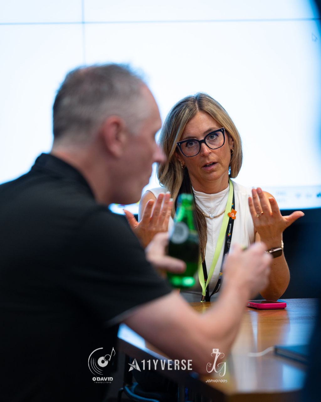 Two people in conversation across a meeting room table, one gesturing with both hands toward the other who listens with a thoughtful expression, with a bottled drink and a phone on the table and a bright screen glowing behind.