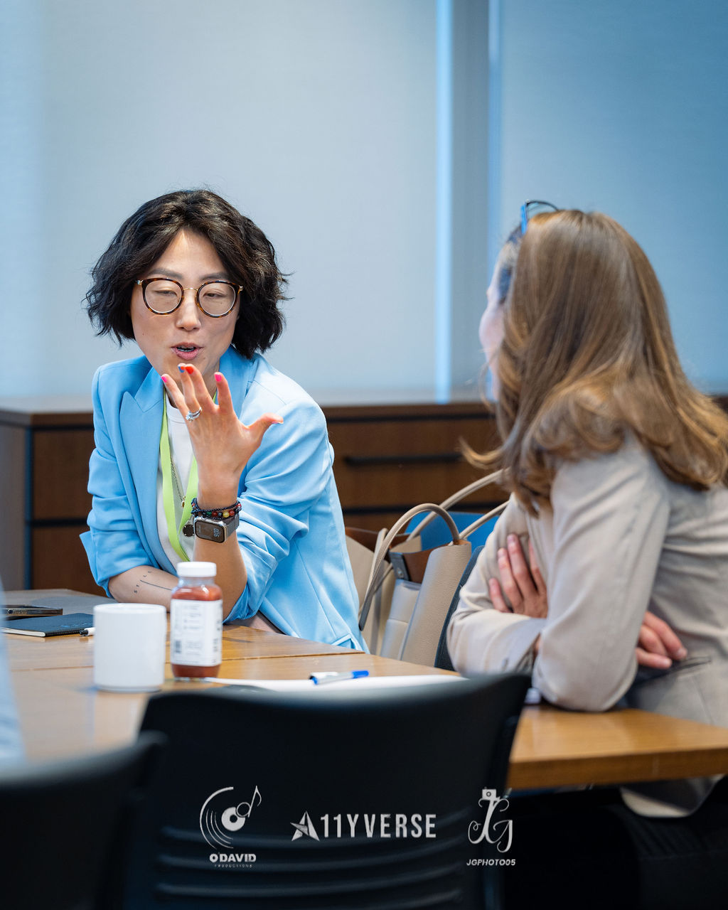 A person in a light blue blazer gestures with one hand while speaking to another seated across a meeting room table, with a coffee cup and a bottled drink in front of them.