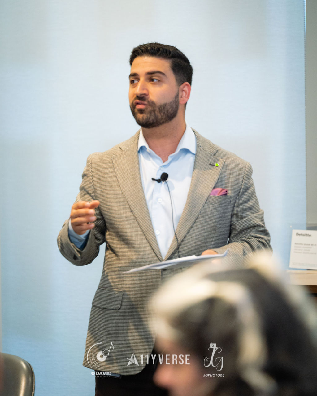 A speaker in a blazer with a clip-on microphone gestures while presenting, holding notes, in front of a plain backdrop.