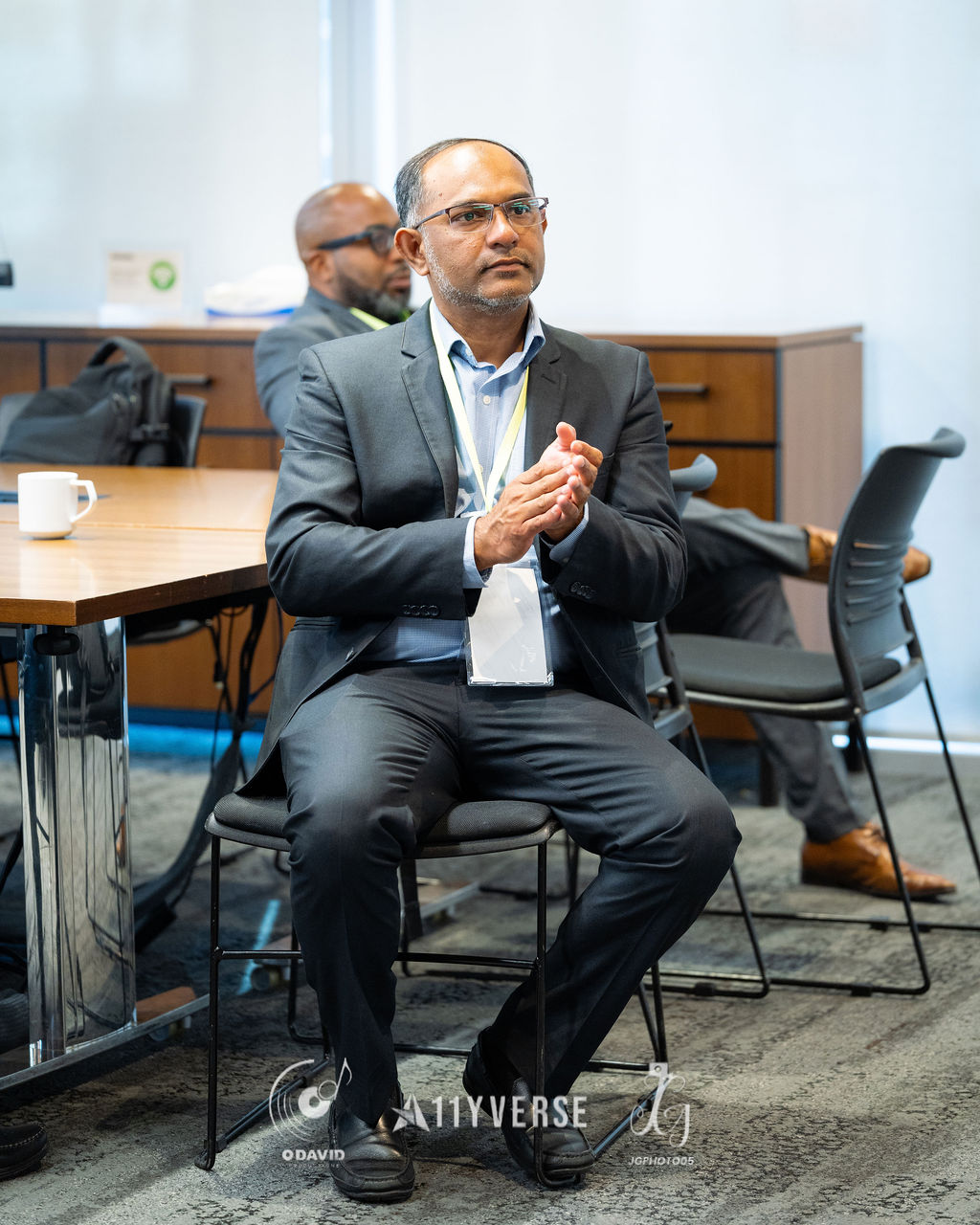 A person in a suit and lanyard sits with hands clasped, listening attentively during a session, with a coffee cup on the table behind.