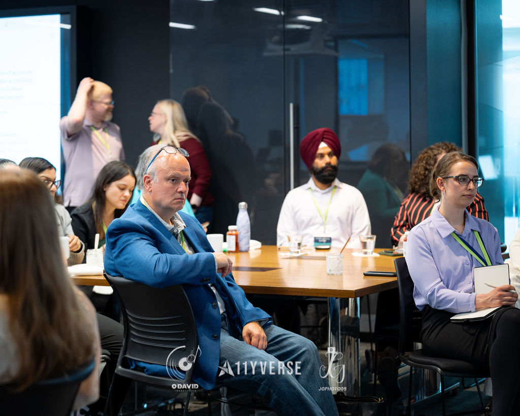 Attendees seated around tables in a glass-walled meeting room listening to a session, some taking notes.