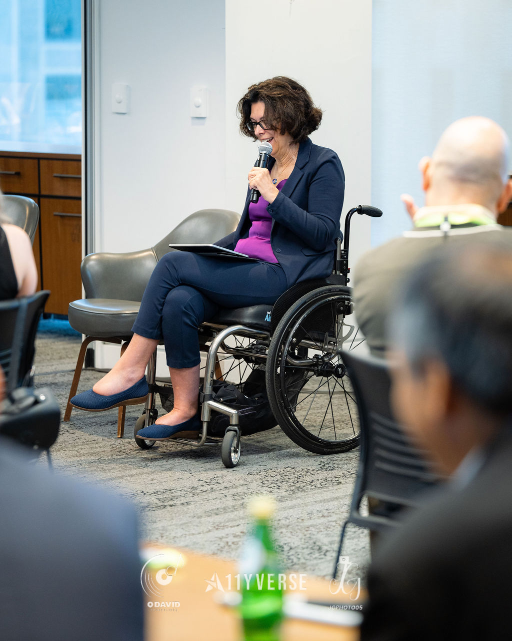 A person seated in a manual wheelchair speaks into a handheld microphone with notes on their lap, smiling, while the audience listens in the foreground.