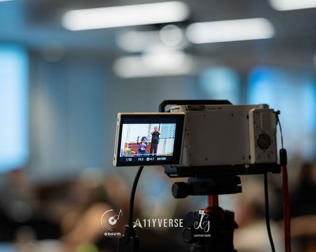 A video camera on a tripod with its flip-out screen showing a speaker being filmed, the session room softly blurred behind.