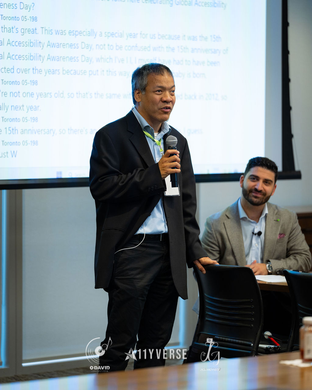 A person stands and speaks into a microphone while another seated beside them smiles, in front of a large screen showing live captions of the conversation.