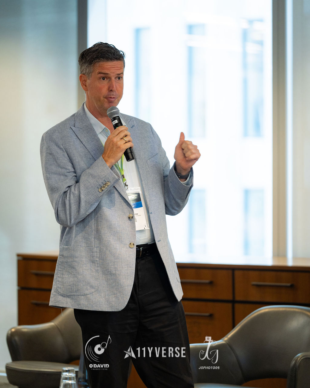 A speaker in a light blazer holds a microphone and gives a thumbs-up while talking, standing near a bright window.