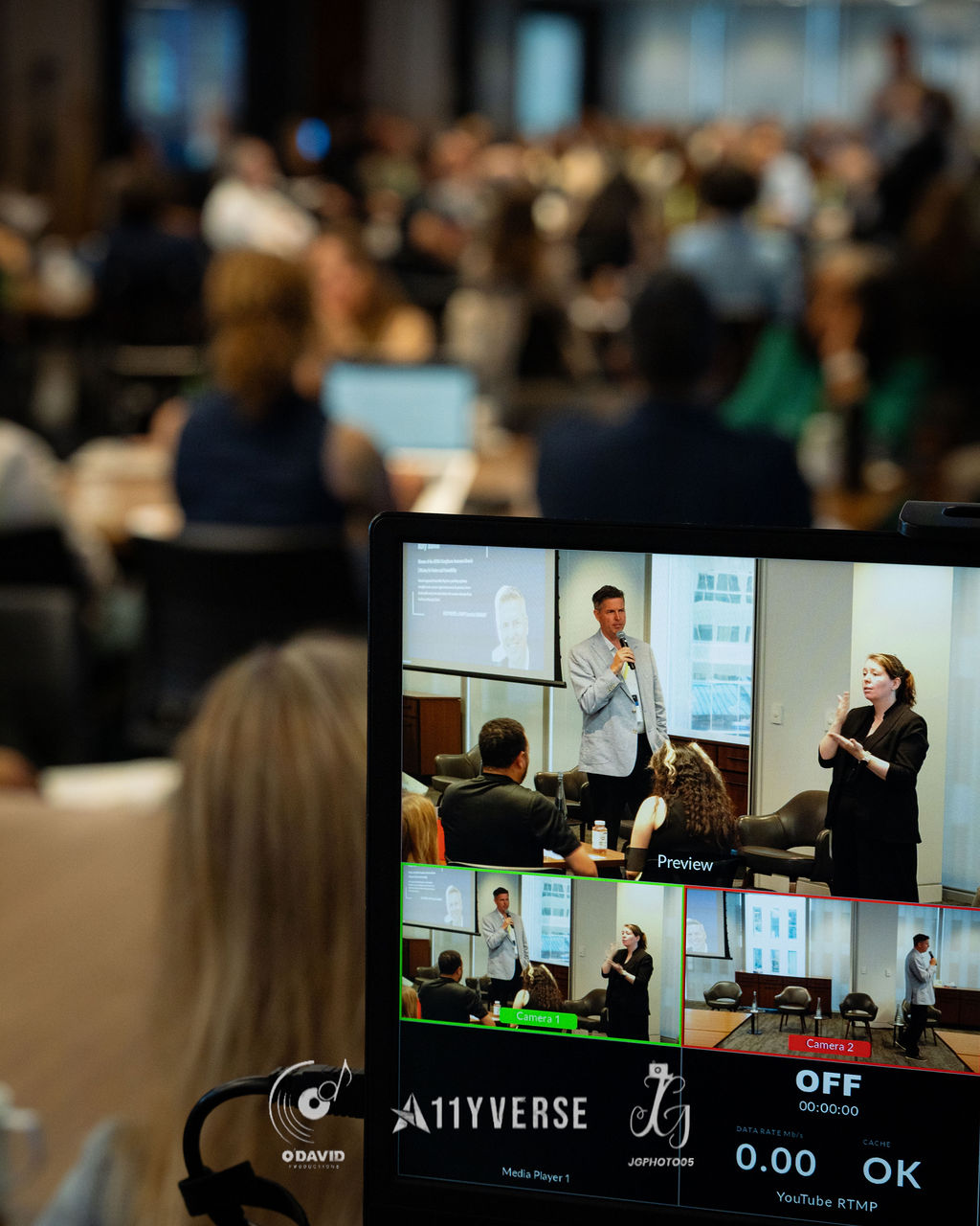 A live-streaming control monitor shows several camera feeds of a speaker and a sign language interpreter, labelled as preview and camera views, with a large audience blurred behind.