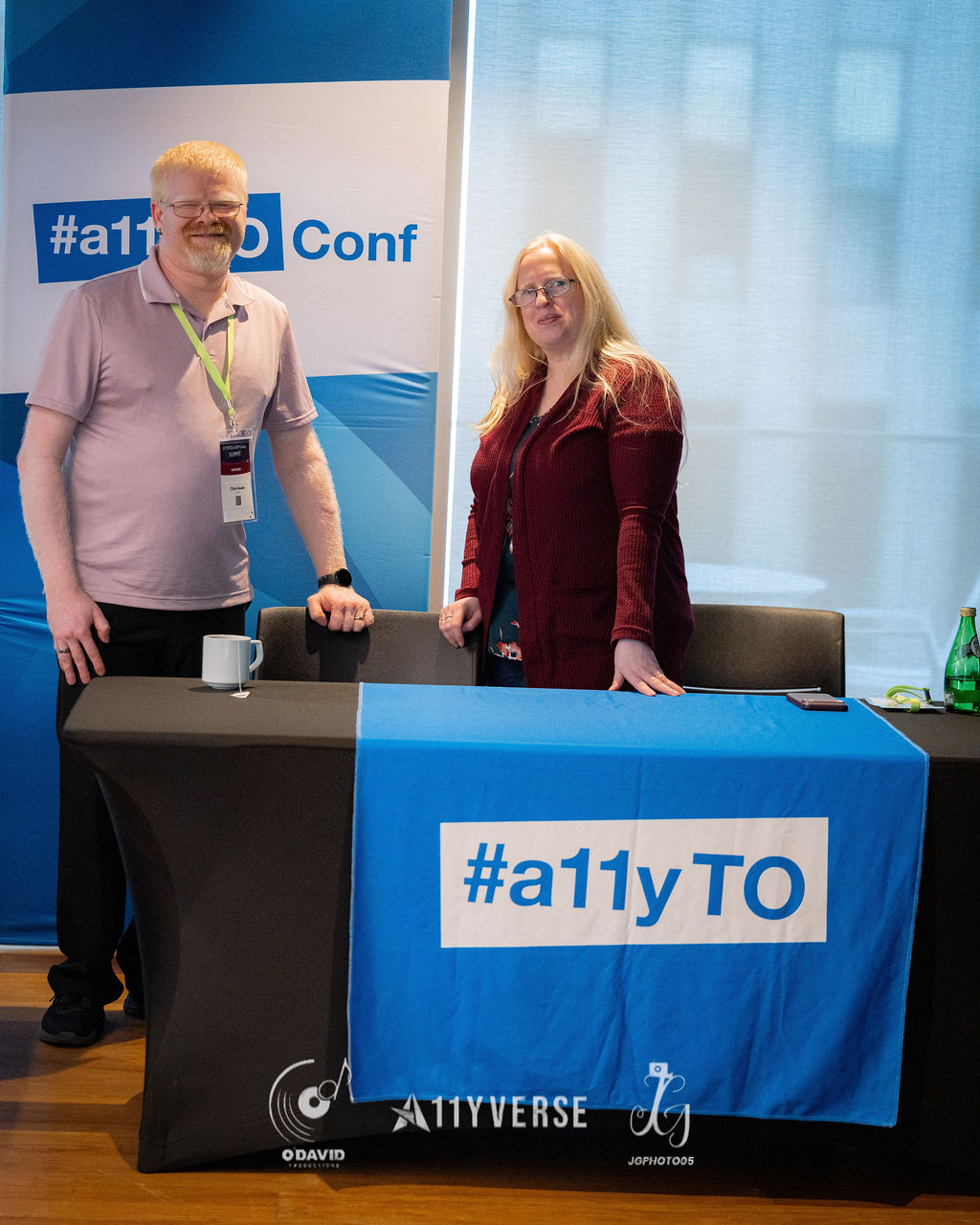 Two people stand smiling behind a welcome desk draped with a blue hashtag a11yTO cloth, in front of a matching conference banner.