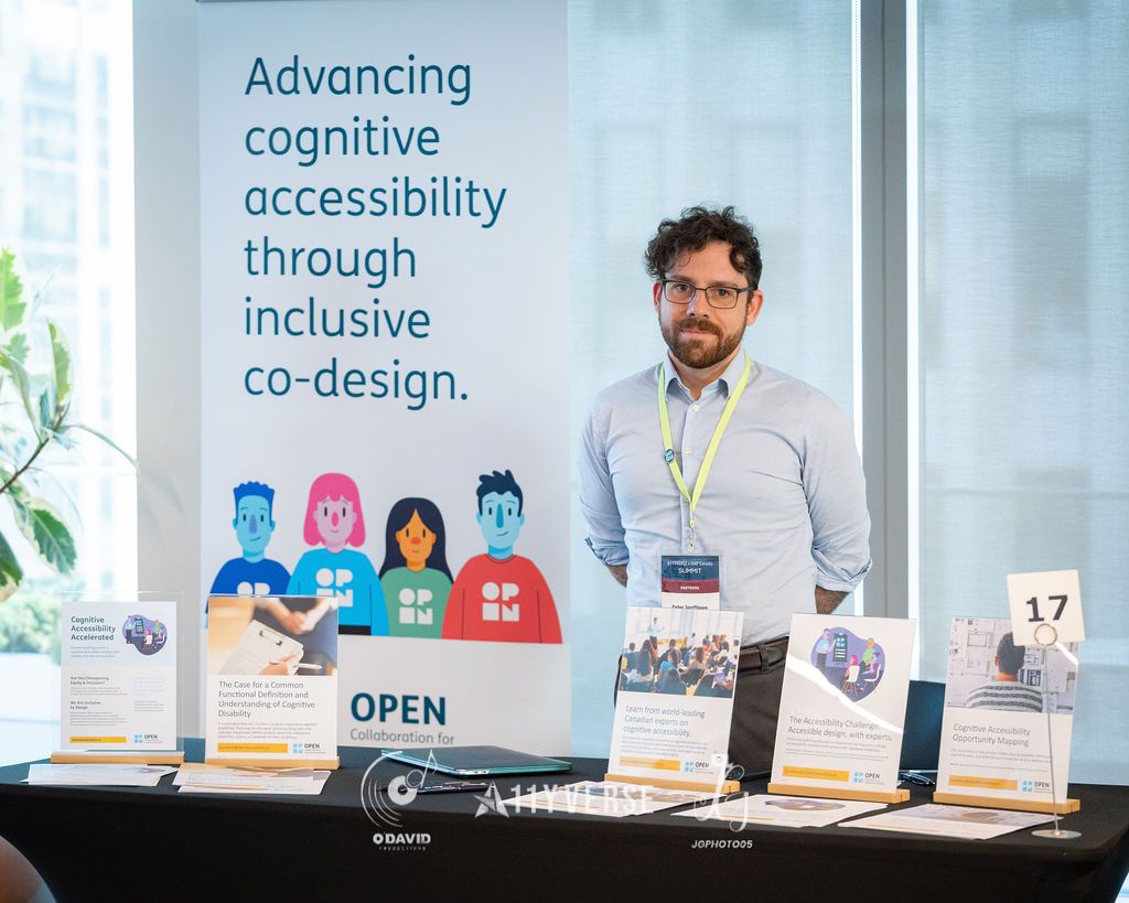 A person stands at the OPEN booth beside a banner about advancing cognitive accessibility through inclusive co-design, with printed handouts and table cards arranged in front.