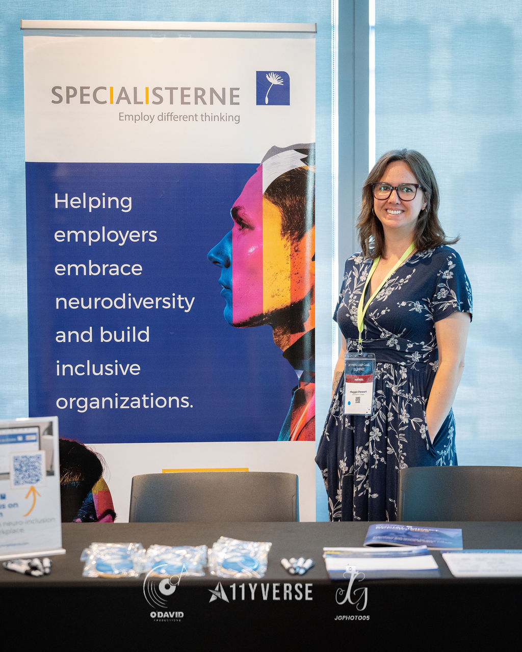 A person stands smiling at the Specialisterne booth beside a banner about helping employers embrace neurodiversity and build inclusive organizations, with sample items on the table.