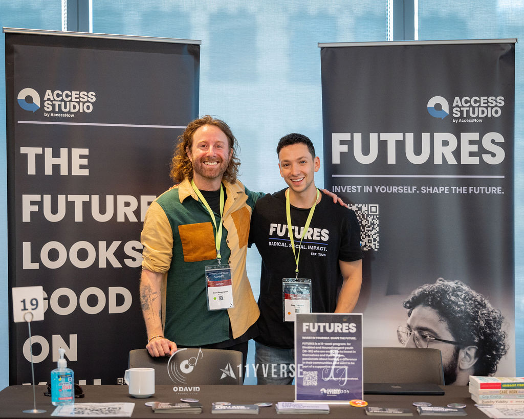 Two people stand arm in arm and smiling behind an exhibitor booth, in front of Access Studio and Futures banners, with hand sanitizer and brochures on the table.