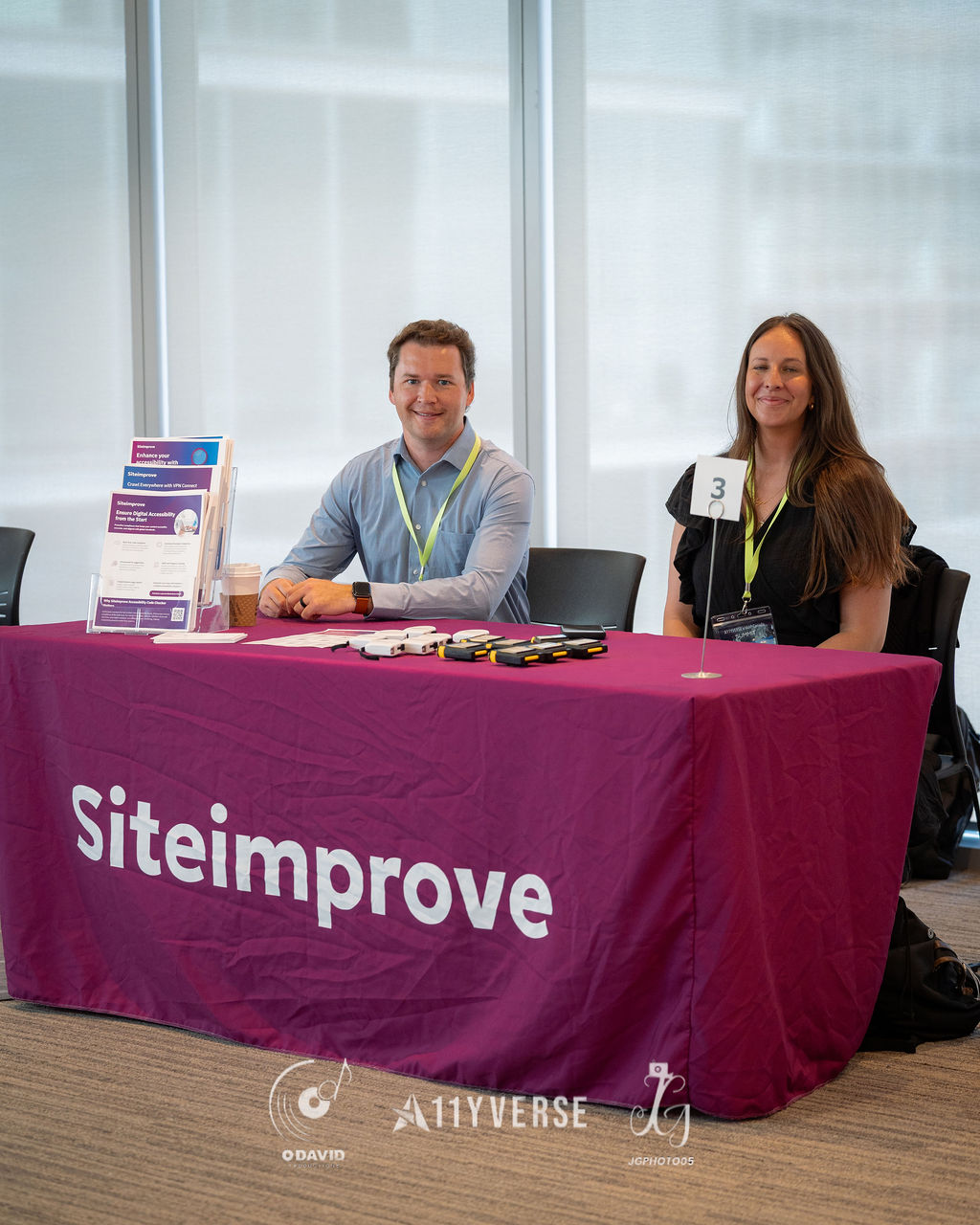 Two people sit and smile behind the Siteimprove booth table, with a brochure stand and printed materials beside them.