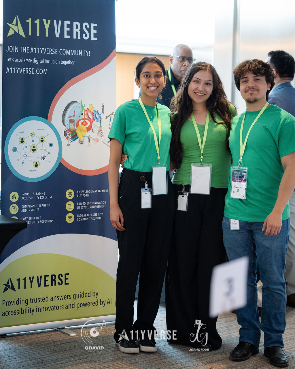 Three people in matching green shirts stand together and smile in front of an A11YVERSE community banner at the booth.