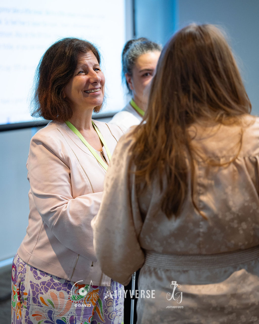 A person in a light blazer smiles warmly while in conversation with another whose back is to the camera, a presentation screen glowing behind.