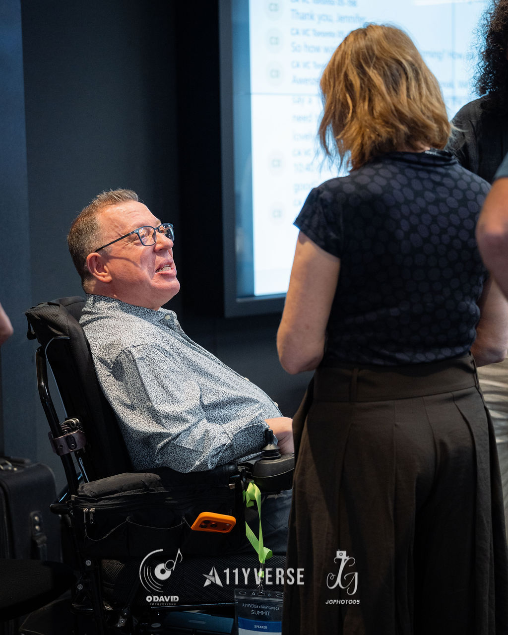 A person seated in a powered wheelchair, badge marked as a speaker, talks with someone standing beside them in front of a captioned presentation screen.