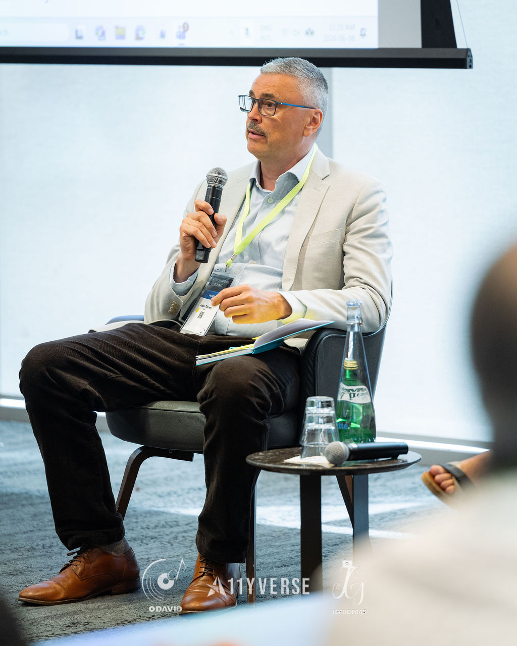 A panelist in a light blazer sits in an armchair speaking into a handheld microphone with notes on their lap, a side table holding water nearby.