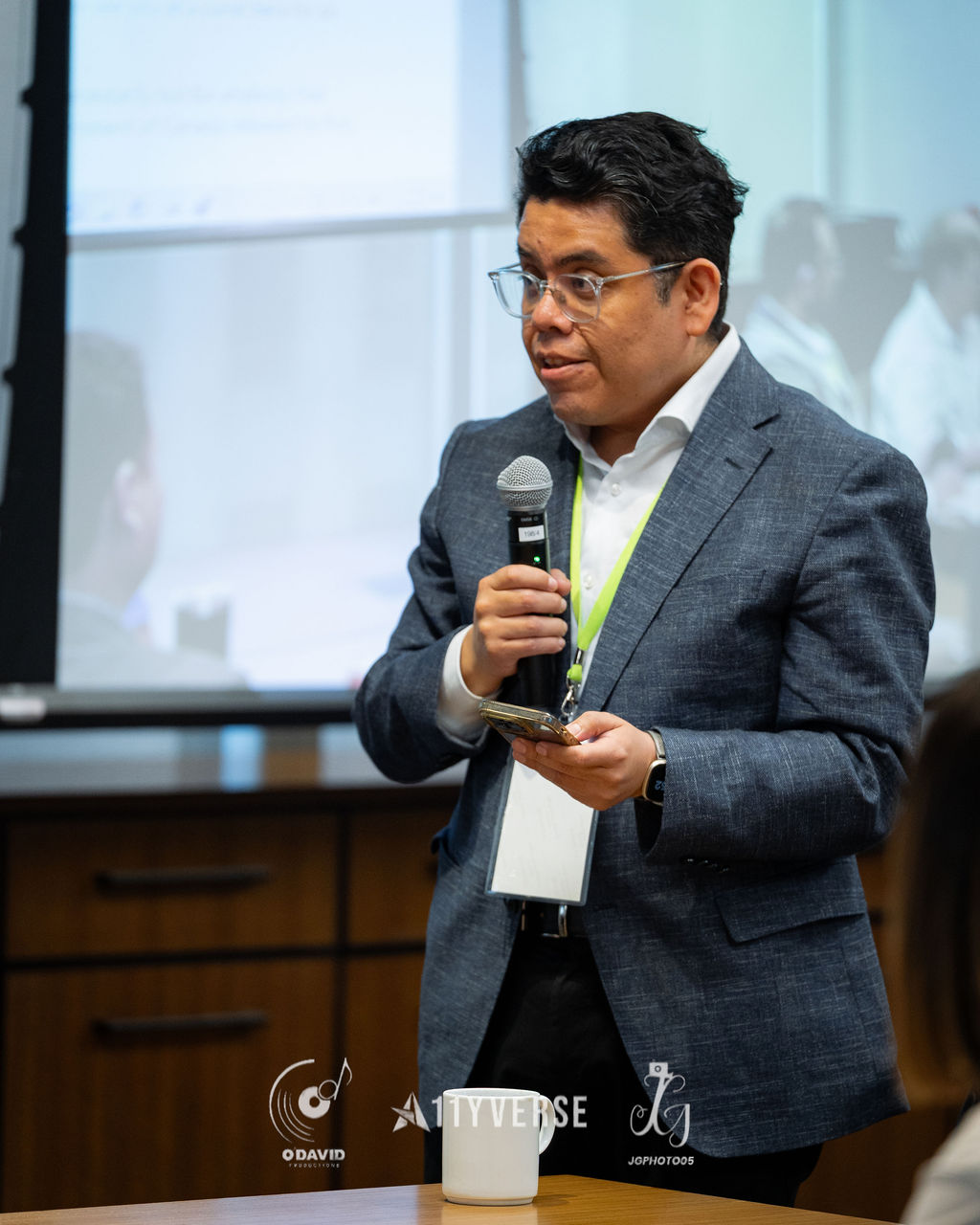 A speaker in a grey blazer holds a microphone and speaks, holding a phone and badge, with a presentation screen behind and a coffee mug in the foreground.