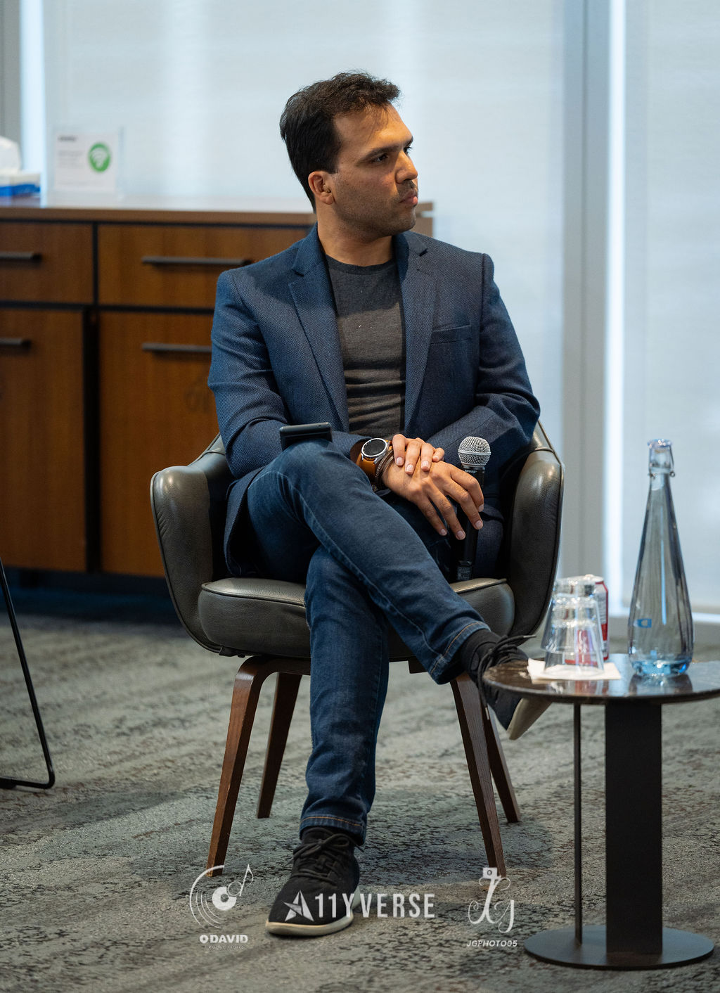 A panelist sits in an armchair resting a handheld microphone on one knee while listening and looking to the side, a side table with water nearby.