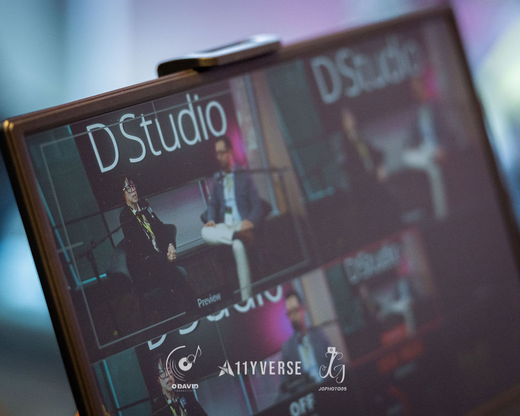 A monitor displays a multi-camera recording layout labelled D Studio, showing two seated panelists in the preview pane, with the venue blurred behind.