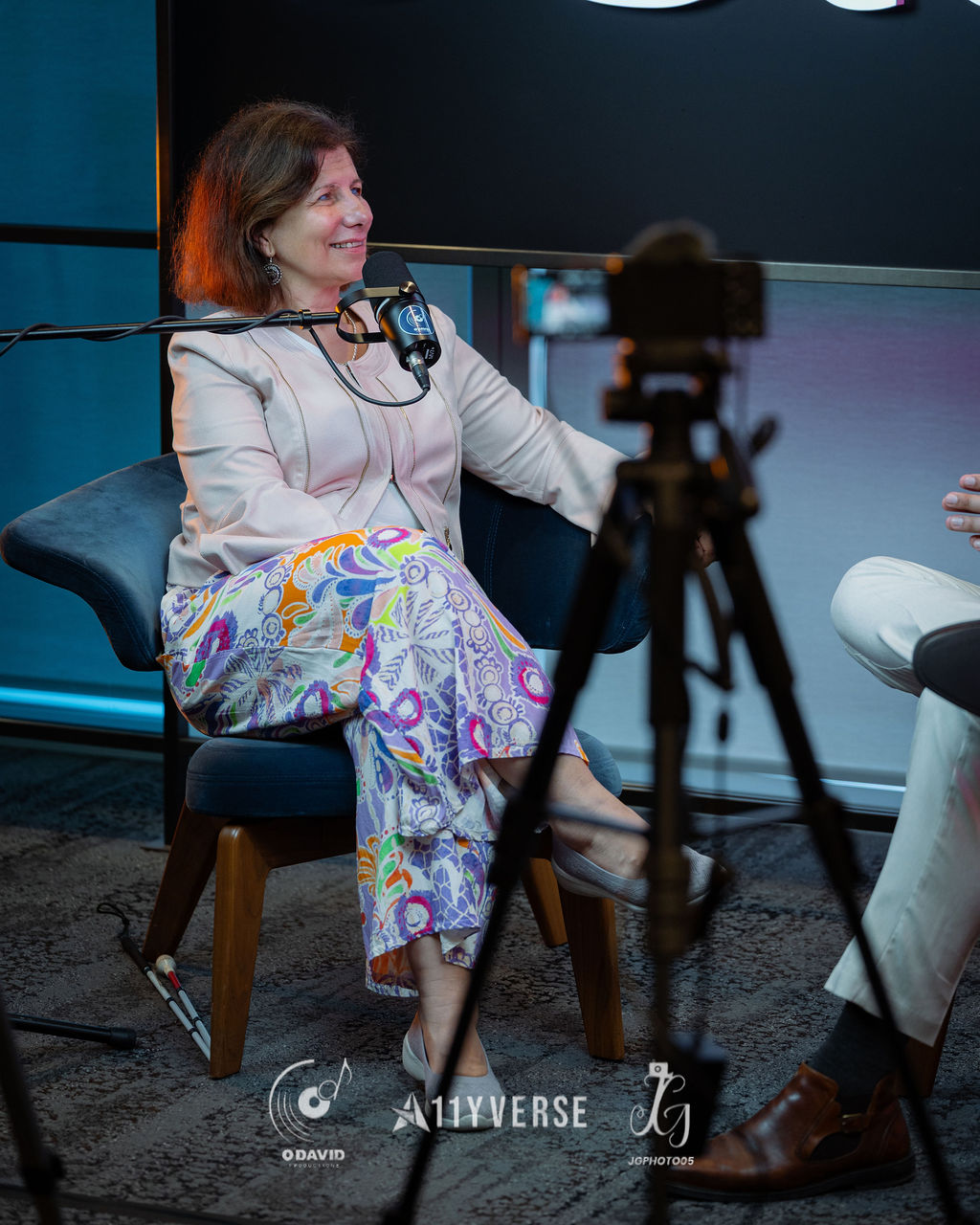 A person sits in an armchair smiling during a recorded interview, speaking into a podcast microphone, with a white cane resting against the chair and a film camera in the foreground.
