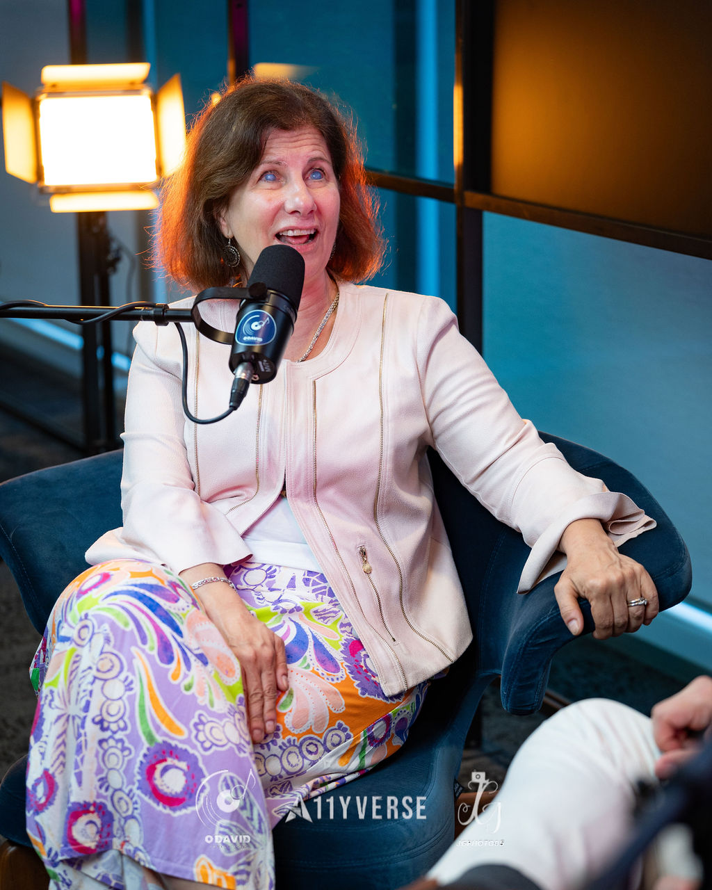 A person sits in an armchair speaking into a podcast microphone during a recorded interview, studio lighting glowing behind.