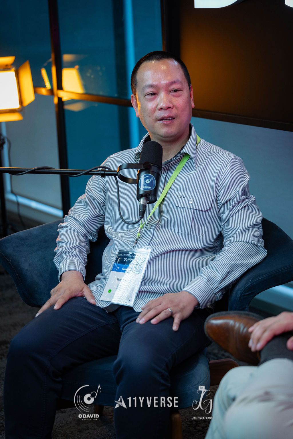 A person sits in an armchair speaking into a podcast microphone during a recorded interview, a lanyard around their neck and studio lights behind.
