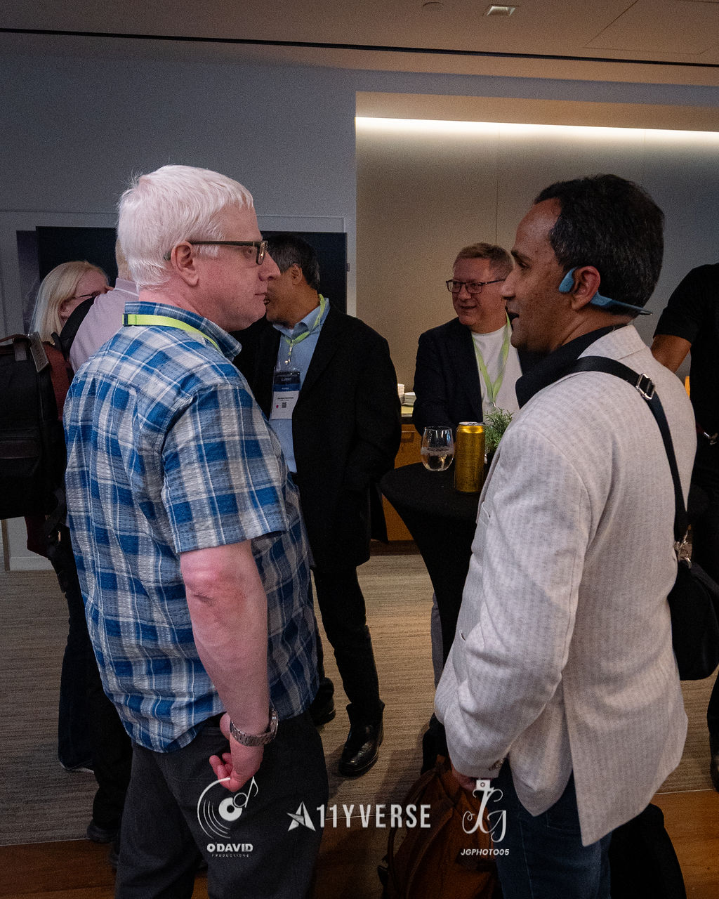 Two attendees stand face to face in conversation during a networking break, with others mingling around a high table behind.
