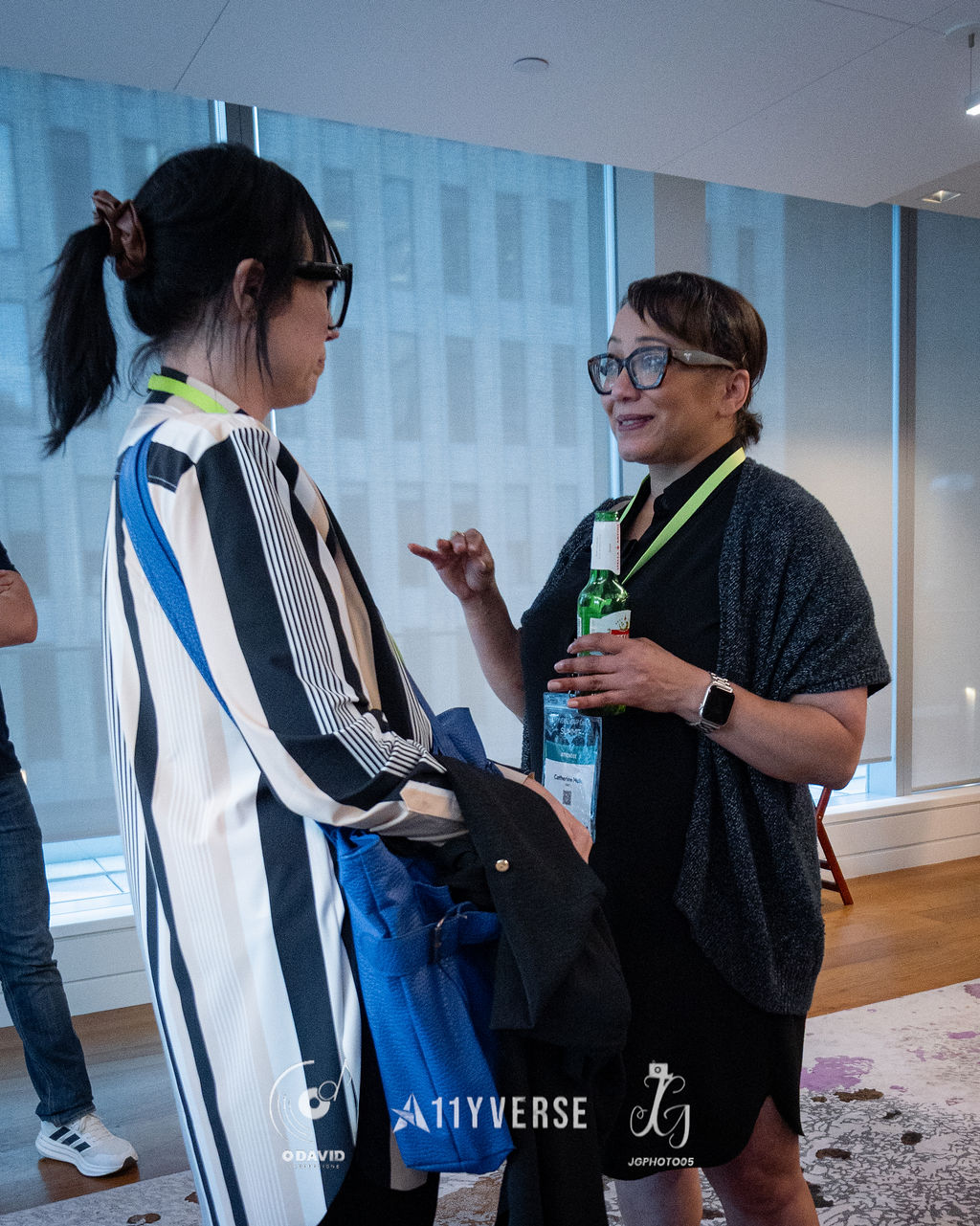 Two attendees stand talking during a break, one holding a tote bag and jacket while the other gestures with a bottled drink, a city view through the window behind.