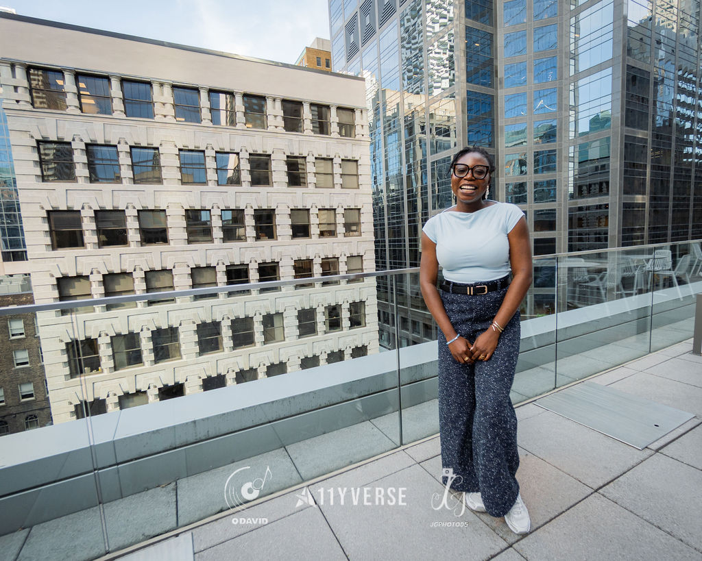 A person stands and smiles on an outdoor rooftop terrace with a glass railing, city buildings behind.