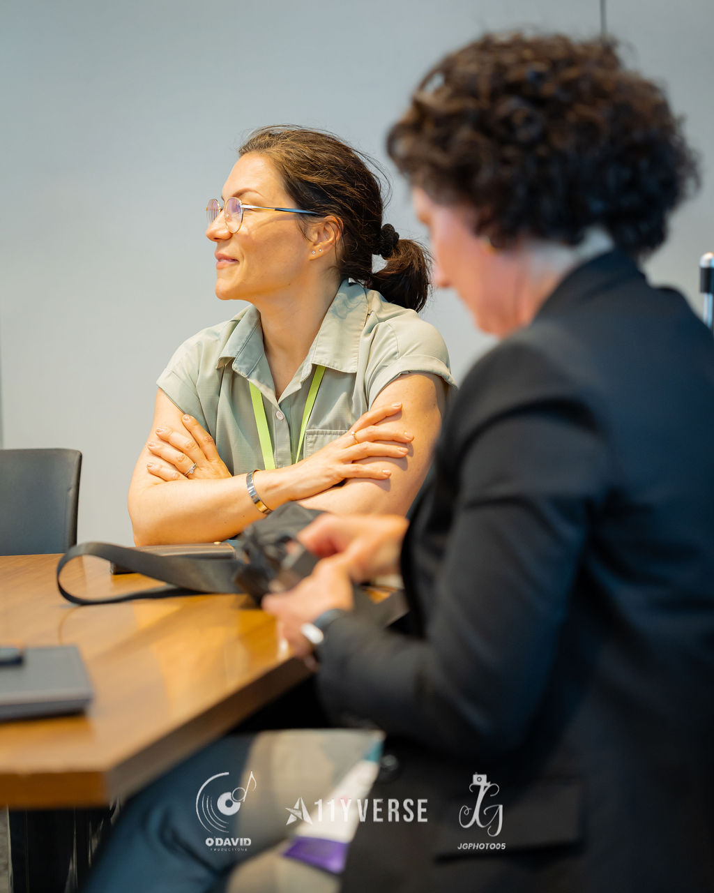 Two people sit at a table in conversation, one with arms crossed listening and smiling while the other, seen from behind, speaks.