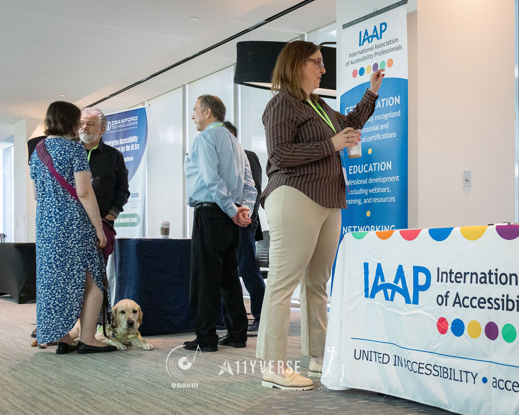 A guide dog in a harness lies calmly on the floor beside its handler at the IAAP booth, with the International Association of Accessibility Professionals banner and table behind, and people standing in conversation.