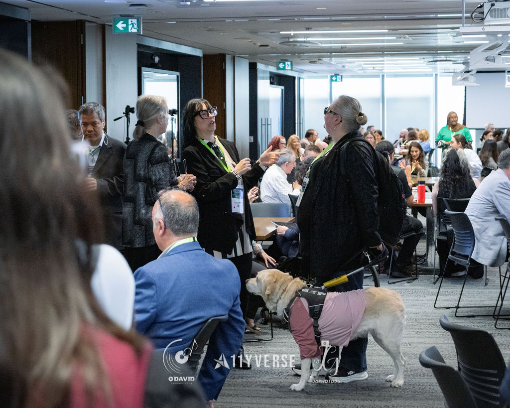 A busy session room with attendees at tables, where a guide dog in a harness and coat stands beside its standing handler in the foreground, an exit sign overhead.